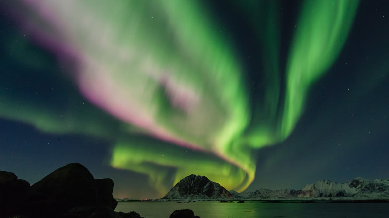 Green and pink aurora swirling over snowy peaks and a calm bay in Lofoten, Northern Norway.