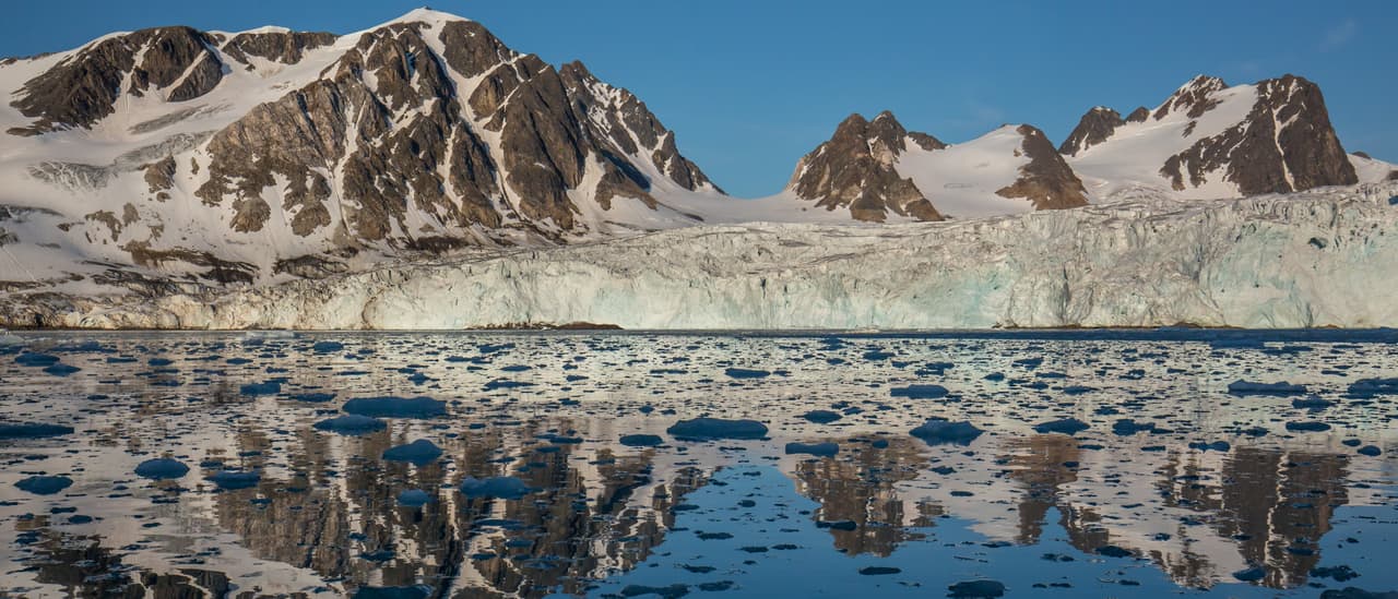 A glacier front below snowy peaks, mirrored in a calm fjord scattered with floating ice, in north-west Spitsbergen, Svalbard.