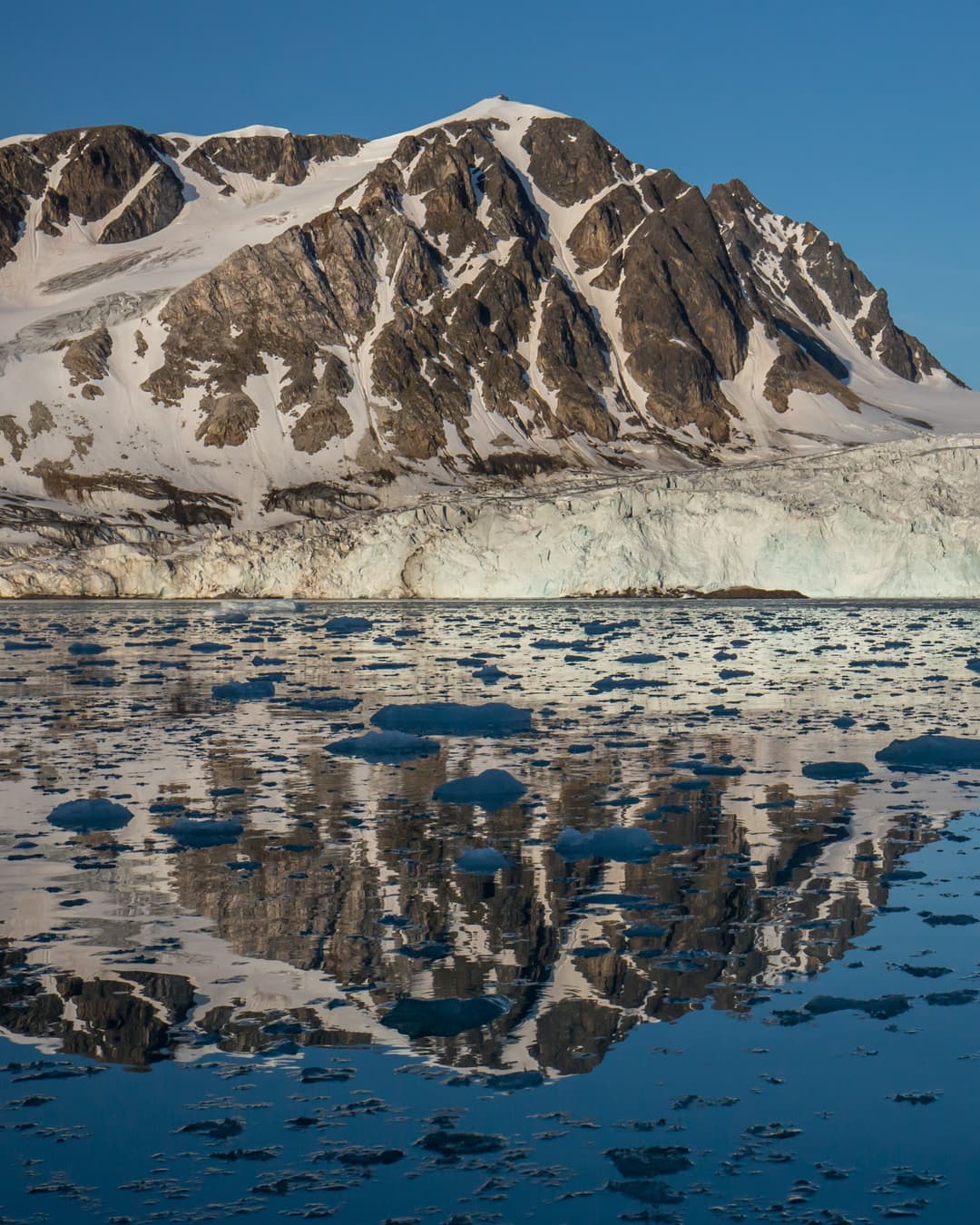 A glacier front below snowy peaks, mirrored in a calm fjord scattered with floating ice, in north-west Spitsbergen, Svalbard.